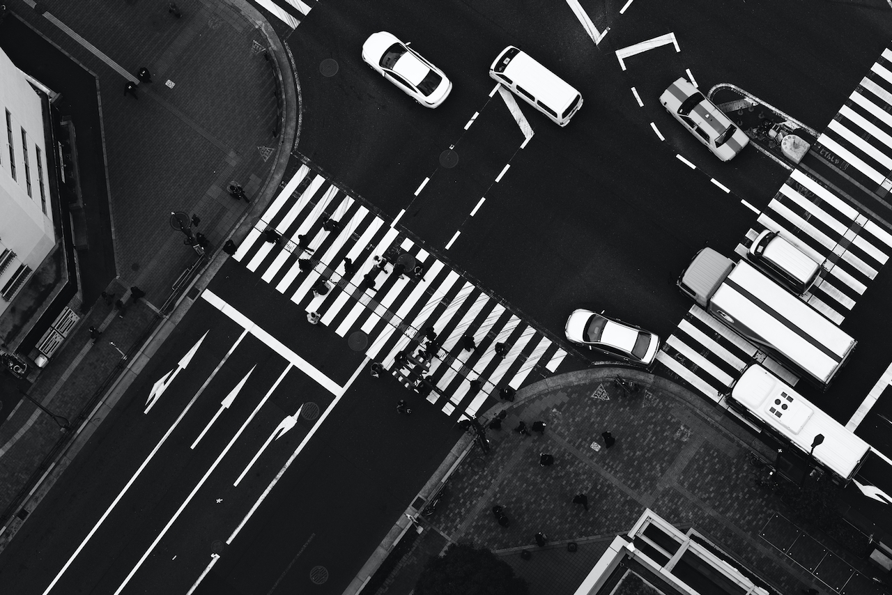 Cars representing organic traffic turn at an intersection while pedestrians use a crosswalk.